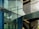 A modern architectural scene featuring a glass facade with visible reflections and metallic frames. The scene includes an internal structure with stairs and metal railings, casting shadows across the glass surface.