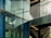 A modern architectural scene featuring a glass facade with visible reflections and metallic frames. The scene includes an internal structure with stairs and metal railings, casting shadows across the glass surface.
