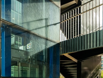 A modern architectural scene featuring a glass facade with visible reflections and metallic frames. The scene includes an internal structure with stairs and metal railings, casting shadows across the glass surface.