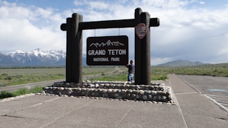 A wooden sign for Grand Teton National Park stands on a stone base. In the background, there are snow-capped mountains under a partly cloudy sky. A person stands near the sign, looking towards it. The surrounding area includes an asphalt road and a grassy landscape.