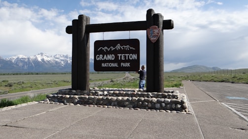 A wooden sign for Grand Teton National Park stands on a stone base. In the background, there are snow-capped mountains under a partly cloudy sky. A person stands near the sign, looking towards it. The surrounding area includes an asphalt road and a grassy landscape.