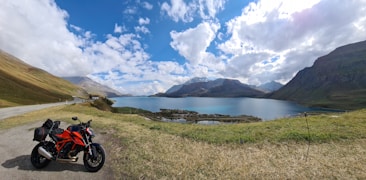 A scenic landscape featuring a vibrant red and black motorcycle parked on a roadside. The scene includes a clear blue lake surrounded by mountains under a sky with scattered clouds. The grassy terrain and winding road suggest a peaceful and adventurous setting.