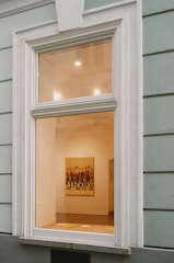 A large window on a building wall reveals the interior of an art gallery, featuring a brightly lit room with a painting of several people walking. The walls are painted a soft cream color, and some lighting fixtures are visible on the ceiling, casting light onto the artwork.