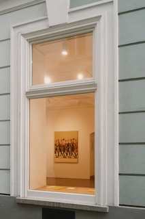 A large window on a building wall reveals the interior of an art gallery, featuring a brightly lit room with a painting of several people walking. The walls are painted a soft cream color, and some lighting fixtures are visible on the ceiling, casting light onto the artwork.