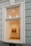 A large window on a building wall reveals the interior of an art gallery, featuring a brightly lit room with a painting of several people walking. The walls are painted a soft cream color, and some lighting fixtures are visible on the ceiling, casting light onto the artwork.
