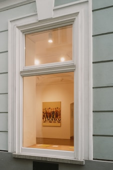 A large window on a building wall reveals the interior of an art gallery, featuring a brightly lit room with a painting of several people walking. The walls are painted a soft cream color, and some lighting fixtures are visible on the ceiling, casting light onto the artwork.
