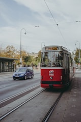 A red tram is traveling along tracks on a city street, with several passengers visible inside. To the left, a blue car drives parallel to the tram. The scene is set against an urban background featuring streetlights and trees lining the road.