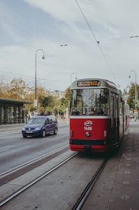A red tram is traveling along tracks on a city street, with several passengers visible inside. To the left, a blue car drives parallel to the tram. The scene is set against an urban background featuring streetlights and trees lining the road.