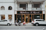 A street view featuring two storefronts with classic architectural details. The first storefront, 'Möbelhaus H. Marek', has an elegant, traditional facade with large windows and black signage with gold lettering. The second storefront, 'werkbank', is modern with a minimalist white sign and large, arched glass windows showcasing interior products. In front of the stores, three vehicles are parked: two white cars and a red moped.