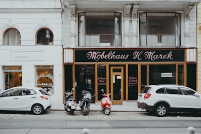A street view featuring two storefronts with classic architectural details. The first storefront, 'Möbelhaus H. Marek', has an elegant, traditional facade with large windows and black signage with gold lettering. The second storefront, 'werkbank', is modern with a minimalist white sign and large, arched glass windows showcasing interior products. In front of the stores, three vehicles are parked: two white cars and a red moped.