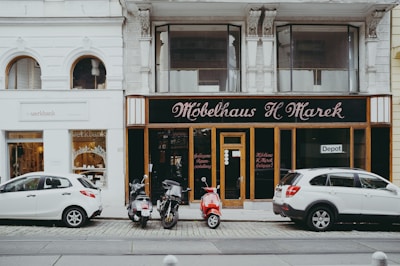 A street view featuring two storefronts with classic architectural details. The first storefront, 'Möbelhaus H. Marek', has an elegant, traditional facade with large windows and black signage with gold lettering. The second storefront, 'werkbank', is modern with a minimalist white sign and large, arched glass windows showcasing interior products. In front of the stores, three vehicles are parked: two white cars and a red moped.
