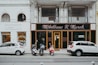 A street view featuring two storefronts with classic architectural details. The first storefront, 'M&ouml;belhaus H. Marek', has an elegant, traditional facade with large windows and black signage with gold lettering. The second storefront, 'werkbank', is modern with a minimalist white sign and large, arched glass windows showcasing interior products. In front of the stores, three vehicles are parked: two white cars and a red moped.