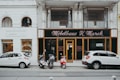 A street view featuring two storefronts with classic architectural details. The first storefront, 'M&ouml;belhaus H. Marek', has an elegant, traditional facade with large windows and black signage with gold lettering. The second storefront, 'werkbank', is modern with a minimalist white sign and large, arched glass windows showcasing interior products. In front of the stores, three vehicles are parked: two white cars and a red moped.
