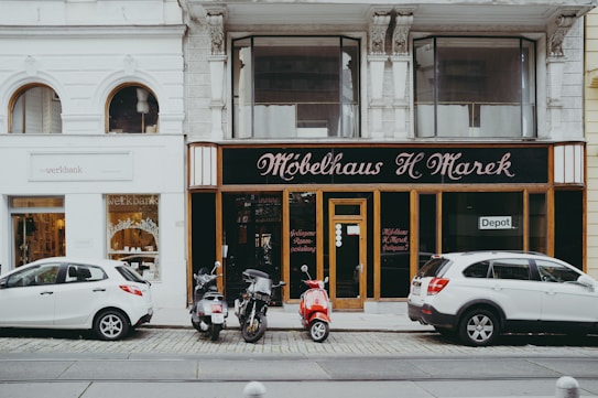 A street view featuring two storefronts with classic architectural details. The first storefront, 'M&ouml;belhaus H. Marek', has an elegant, traditional facade with large windows and black signage with gold lettering. The second storefront, 'werkbank', is modern with a minimalist white sign and large, arched glass windows showcasing interior products. In front of the stores, three vehicles are parked: two white cars and a red moped.