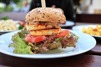 A close-up of a gourmet sandwich placed on a white plate, featuring a sesame seed bun topped with grilled halloumi cheese, a slice of tomato, and crunchy lettuce. In the background, a blurred view shows other plates, including one with french fries, suggesting a casual dining setting.