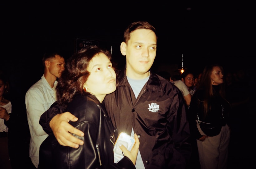 A group of people are in a dimly lit setting, possibly at a social or event venue. A man and a woman are in the foreground; the man is wearing a dark jacket and the woman is in a leather jacket holding a smartphone. They appear to be posing for a photograph, with the man smiling gently and the woman making a playful face. Other people are visible in the background, engaging in conversation.