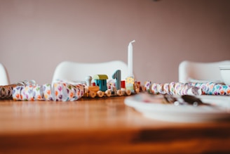 A wooden toy train with a colorful design sits on a table surrounded by a festive paper garland featuring balloon patterns. A white candle is placed on the train, and the background shows white chairs and part of a dining setting with plates and cutlery slightly out of focus.