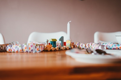 A wooden toy train with a colorful design sits on a table surrounded by a festive paper garland featuring balloon patterns. A white candle is placed on the train, and the background shows white chairs and part of a dining setting with plates and cutlery slightly out of focus.