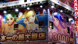 An illuminated building facade is covered with large, colorful anime-style illustrations featuring characters with vibrant hair colors. The artwork includes two female characters and one male character, surrounded by bright and lively aquatic-themed graphics. Japanese text in bold fonts is prominently displayed across the facade.