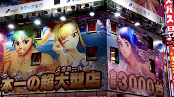 An illuminated building facade is covered with large, colorful anime-style illustrations featuring characters with vibrant hair colors. The artwork includes two female characters and one male character, surrounded by bright and lively aquatic-themed graphics. Japanese text in bold fonts is prominently displayed across the facade.