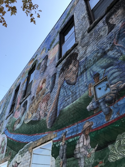 A vibrant mural painted on the side of a building depicts various people engaged in different activities. The artwork includes diverse figures, some holding objects and others immersed in tasks, set against a vivid background.