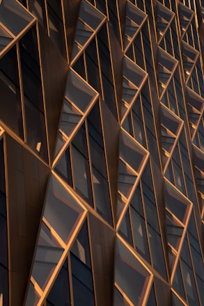 A close-up of a modern architectural facade featuring a series of geometric patterns composed of glass and metal. The facade appears to capture light, giving it a dynamic appearance with reflections and shadows. The design includes diagonal and triangular shapes arranged in a repeating pattern.