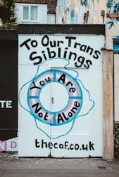 A painted wall with a supportive message for transgender individuals, featuring a circular design with blue and pink hues. The text reads 'To Our Trans Siblings, You Are Not Alone' and includes a website URL at the bottom.