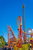 A vibrant amusement park scene featuring a roller coaster with red and yellow tracks. The roller coaster includes steep drops and loops. A tall observation tower stands in the background against a bright blue sky. People are gathered at the entrance and walking around the area, enjoying the attraction.