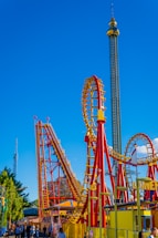 A vibrant amusement park scene featuring a roller coaster with red and yellow tracks. The roller coaster includes steep drops and loops. A tall observation tower stands in the background against a bright blue sky. People are gathered at the entrance and walking around the area, enjoying the attraction.