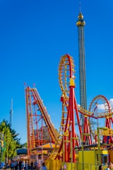 A vibrant amusement park scene featuring a roller coaster with red and yellow tracks. The roller coaster includes steep drops and loops. A tall observation tower stands in the background against a bright blue sky. People are gathered at the entrance and walking around the area, enjoying the attraction.