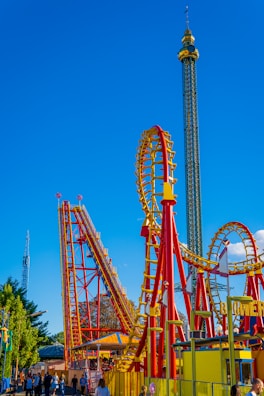 A vibrant amusement park scene featuring a roller coaster with red and yellow tracks. The roller coaster includes steep drops and loops. A tall observation tower stands in the background against a bright blue sky. People are gathered at the entrance and walking around the area, enjoying the attraction.
