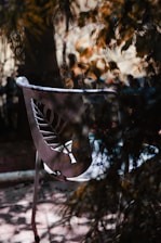 A metal chair with a leaf design cutout in the backrest, placed outdoors. The scene is dappled with light coming through the foliage, creating patterns of shadows and sunlight on the ground and the chair.