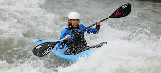 A person is kayaking through turbulent whitewater rapids, wearing a helmet and protective gear. The kayak is blue, and they are holding a paddle with both hands.