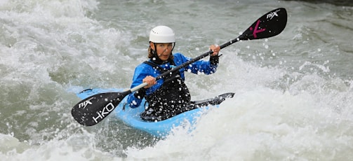 A person is kayaking through turbulent whitewater rapids, wearing a helmet and protective gear. The kayak is blue, and they are holding a paddle with both hands.