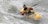 A person is kayaking through turbulent white water rapids. The individual is wearing a helmet and a red, yellow, and black life jacket. The kayak is yellow with a visible brand logo on it. Water splashes around as they navigate through the rapid.