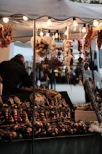 A market stall adorned with decorative hanging items and bright string lights. The stall features a variety of dried floral arrangements and crafted ornaments, with heart-shaped designs prominently displayed. A person stands nearby, partially visible and seemingly engaged with the merchandise.