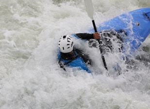 A person wearing a helmet is engaged in white-water kayaking, surrounded by splashing and foaming water. The kayak is blue, and the paddler is gripping the paddle firmly, navigating through the rough waters.