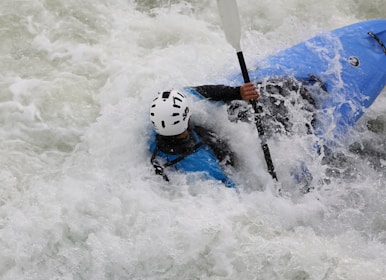 A person wearing a helmet is engaged in white-water kayaking, surrounded by splashing and foaming water. The kayak is blue, and the paddler is gripping the paddle firmly, navigating through the rough waters.