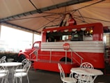 A vintage-style red food truck with Coca-Cola branding, featuring a classic advertisement of a woman holding a Coke bottle. The truck is under a large, open-sided canopy, with white metal tables and chairs set up in front of it. The truck has a retro design with a window for serving customers and displays drinks and snacks inside.