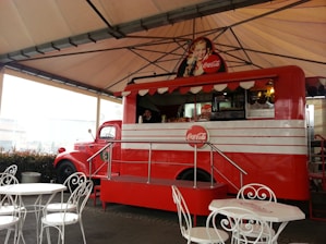A vintage-style red food truck with Coca-Cola branding, featuring a classic advertisement of a woman holding a Coke bottle. The truck is under a large, open-sided canopy, with white metal tables and chairs set up in front of it. The truck has a retro design with a window for serving customers and displays drinks and snacks inside.