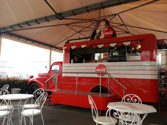 A vintage-style red food truck with Coca-Cola branding, featuring a classic advertisement of a woman holding a Coke bottle. The truck is under a large, open-sided canopy, with white metal tables and chairs set up in front of it. The truck has a retro design with a window for serving customers and displays drinks and snacks inside.