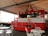 A vintage-style red food truck with Coca-Cola branding, featuring a classic advertisement of a woman holding a Coke bottle. The truck is under a large, open-sided canopy, with white metal tables and chairs set up in front of it. The truck has a retro design with a window for serving customers and displays drinks and snacks inside.