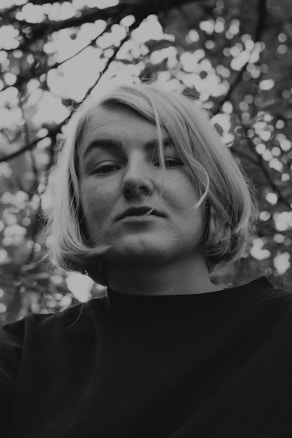 A black and white portrait of a person with short, light-colored hair, wearing a dark top. The person is looking directly at the camera with a neutral expression. The background consists of out-of-focus tree branches and leaves, creating a soft, natural setting.