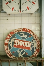 A vintage sports emblem with Cyrillic text features stylized figures in motion, surrounded by red and blue colors. Above it is an old wall clock with faded red and black details on a white tiled background.
