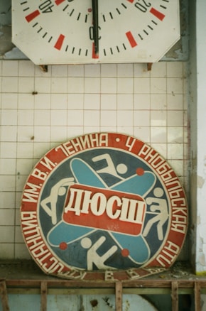 A vintage sports emblem with Cyrillic text features stylized figures in motion, surrounded by red and blue colors. Above it is an old wall clock with faded red and black details on a white tiled background.