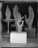 Several large hand sculptures are displayed in a studio setting. The hands are in different gestures, including one in an 'OK' sign. They are placed on pedestals and tables of varying heights, set against a dark backdrop.