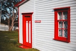 A quaint white house with bright red accents on the door and window frames, featuring a small sign with 'The Cribbies' beside the door. The house is set in a lush green yard with a white picket fence and a tree visible in the background.