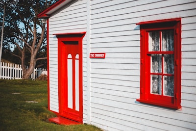 A quaint white house with bright red accents on the door and window frames, featuring a small sign with 'The Cribbies' beside the door. The house is set in a lush green yard with a white picket fence and a tree visible in the background.