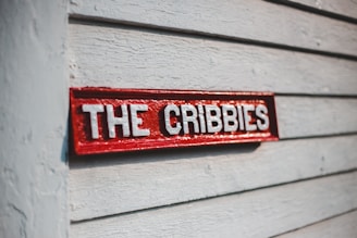 A red wooden sign with white letters spelling 'THE CRIBBIES' is mounted on a gray textured wooden wall. The sunlight casts a shadow, giving depth to the sign's engraved letters.