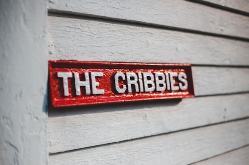 A red wooden sign with white letters spelling 'THE CRIBBIES' is mounted on a gray textured wooden wall. The sunlight casts a shadow, giving depth to the sign's engraved letters.